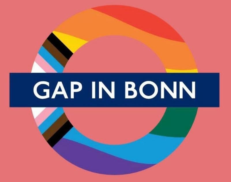 Das Logo erinnert an U-Bahn Symbole. Auf rötlichem Hintergrund ist ein Ring in den Farben der Progress Pride Flag zu sehen. Horizontal queer darüber ist ein blauer Balken mit dem Text "GAP IN BONN" in weiß.
