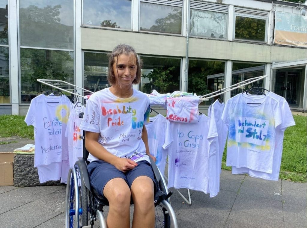 Lena (sie | dey)
Lena sitzt im Rollstuhl vor dem Wäscheständer, an dem wir bei unserer Demonstration die T-Shirts aufgehangen haben. 
Sie hat das Disability Pride Bonn T-Shirt an und lächelt.