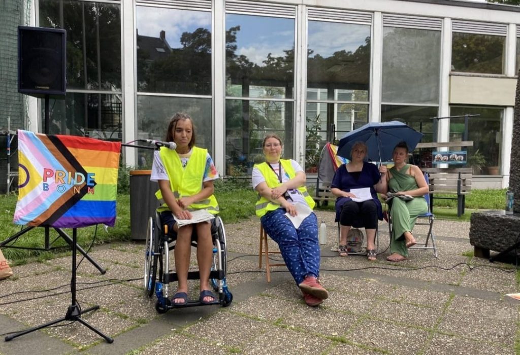 Lena and Solveïg at the microphone
both wearing high-visibility waistcoats.
Lena is sitting in a wheelchair, Solveïg on a folding chair.