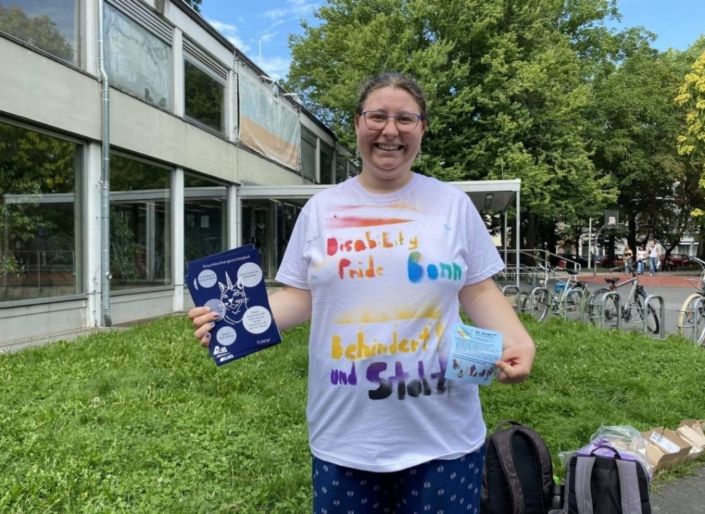 Solveïg (hen | hens)
non•binary, with flyers in hand. Hen is wearing the Disability Pride T-Shirt, which also says "Disabled and Proud" (in the colours of the non•binary pride flag).
