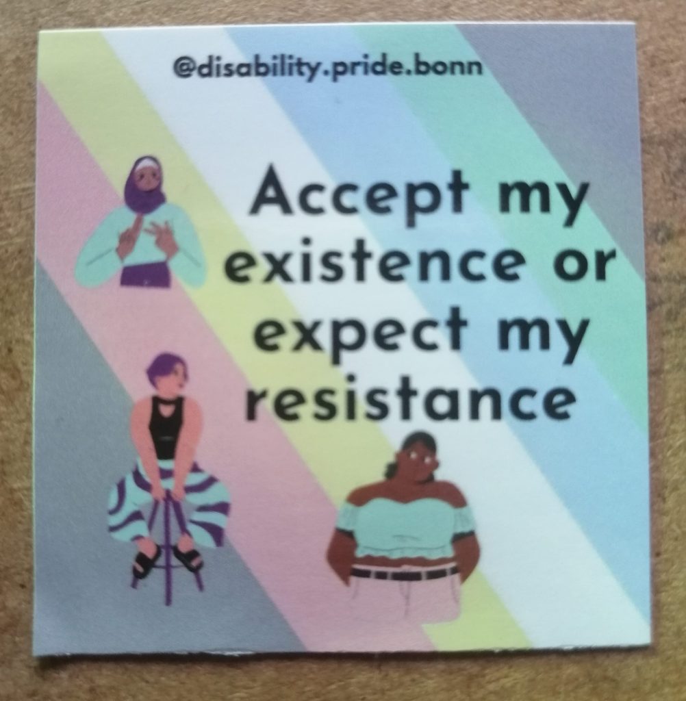 @disability.pride.bonn Accept my existence or expect my resistance. Im Hintergrund ist die Disability Pride Flagge. Davor sind noch 3 Personen. Eine Person mit Kopftuch gebärdet. Eine Person sitzt auf einem Hocker. Eine schwarze, dicke Person halt die Arme hinter den Körper.