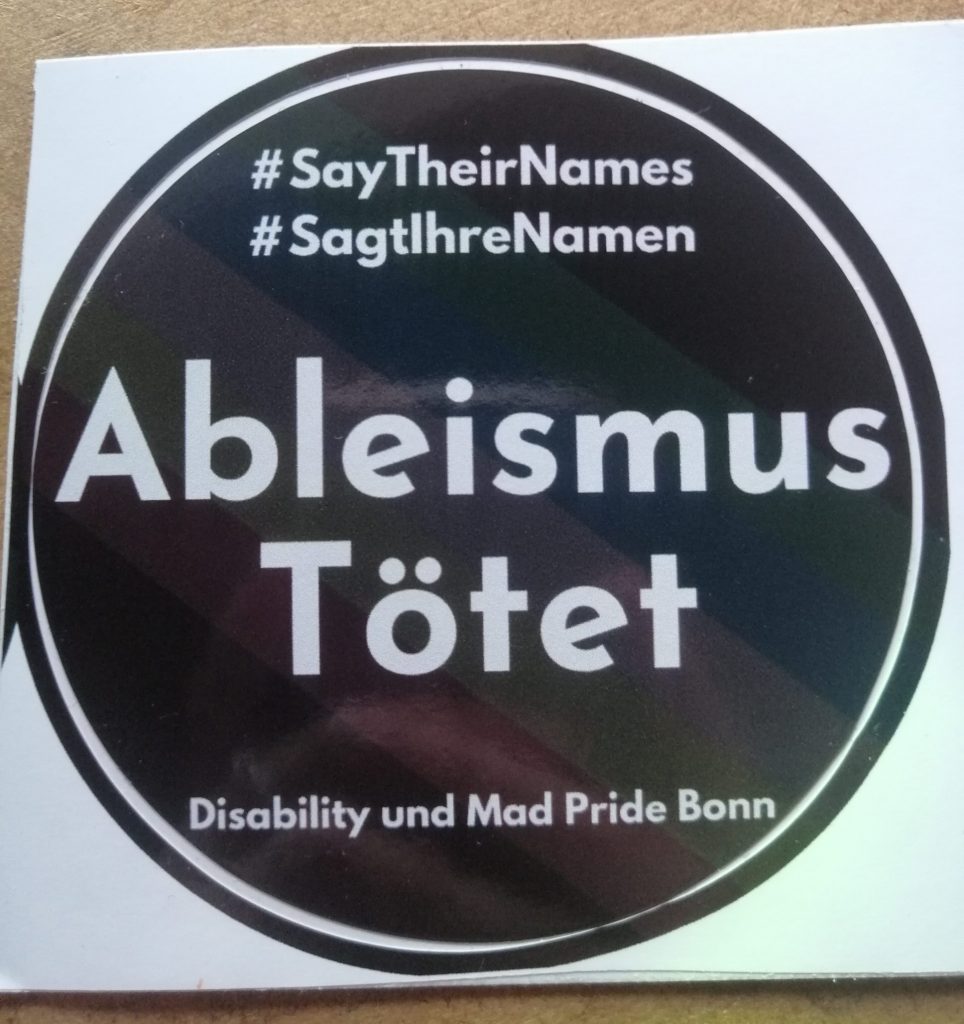 #SayTheirNames #SagtIhreNamen Ableismus Tötet. Disability und Mad Pride Bonn. Der Hintergrund ist schwarz. Ganz leicht ist die Disability Pride Fahne zu sehen.