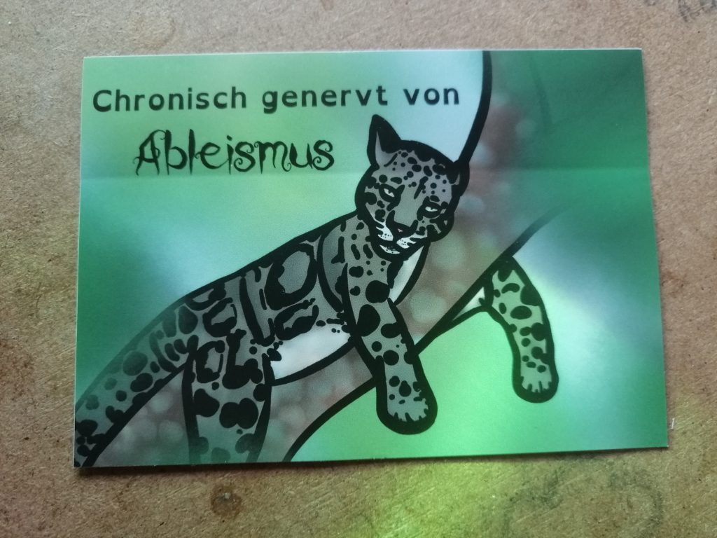 Ein Nebelparder liegt auf einem Ast. Das ist eine Wildkatze. Sie guckt genervt. Text: Chronisch genervt von Ableismus. Der Hintergrund ist grün.