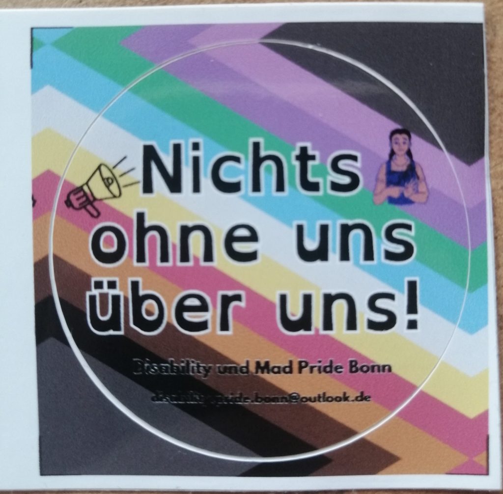 Nichts ohne uns über uns! Disability und Mad Pride Bonn. disability.pride.bonn@outlook.de Im Hintergrund ist die CIMND* Pride Fahne. Die hat einen grauen Hintergrund und schiefe Streifen von links unten nach rechts oben. Die Farben der Streifen sind: schwarz, braun, orange, rot, gelb, weiß, blau, grün, lila, pink. Links ist ein Megafon. Rechts ist eine gebärdende Person.