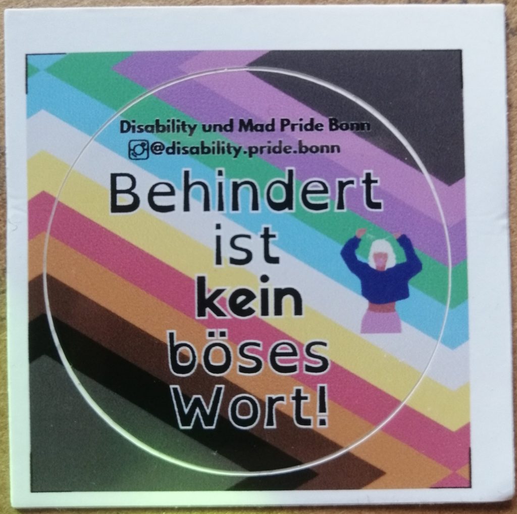 Zu sehen ist ein Sticker der Disability und Mad Pride Bonn. Da steht "Behindert ist kein böses Wort" drauf. Im Hintergrund ist die Disability Pride Fahne, die Koi Katha und Lena entworfen haben. Die Fahne hat einen grauen Hintergrund und schiefe Streifen von links oben nach rechts unten. Die Farben der Streifen sind: hell-lila, dunkel-lila, türkis, blau, weiß, gelb, rot, orange, braun, schwarz.