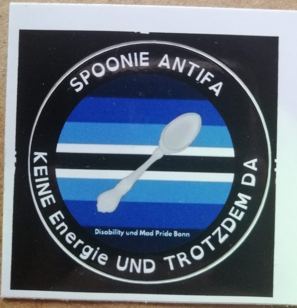 In der Mitte ist die Spoonie Pride Fahne mit einem Löffel. Drum herum steht auf schwarzem Hintergrund: Spoonie Antifa - Keine Energie und trotzdem da. Auf der Flagge steht noch: Disability und Mad Pride Bonn.