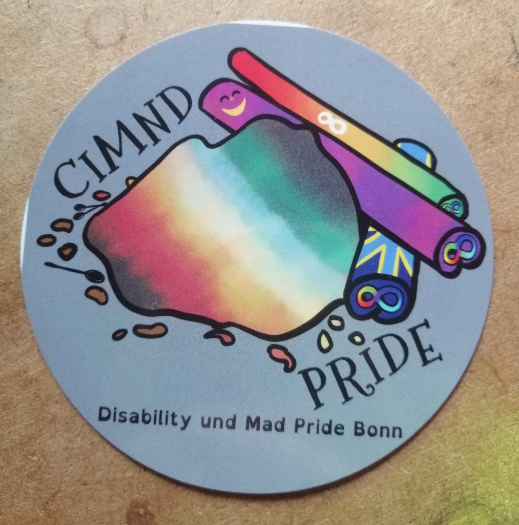 CIMND Pride. Zu sehen sind ein Zimthaufen und mehrere Zimtstangen mit verschiedenen Pride Fahnen: Disability Pride Fahne, Mad Pride, Autistic Pride, Deaf Pride und die Neurodivergenz Schleife und Spoonie Löffel. Disability und Mad Pride Bonn. Der Hintergrund ist grau.