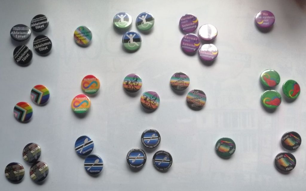Button Designs 2025: CIMND* Schmetterling, Ableismus Tötet #SayTheirNames #SagtIhreNamen, Spoonie, Disability inclusive Inter Progress Pride Flagge, Behindert und Stolz mit Disability/CIMND* Pride Fahne, Actually Autistic mit Autistic Pride Fahne, CIMND* Pride Zimthaufen mit petrol Hintergrund, CIMND* Pride Zimthaufen mit dunkelblauem Hintergrund, Verrückt nach Selbstbestimmung mit Mad Pride Fahne und Grinsen, Disability und Mad Pride Bonn Logo, Ambulatory Wheelchair Users Exist mit der Pride Fahne, Böse Behinderte mit fauchender Katze mit Böse Behinderte Pride Fahne, Spoonie Antifa Keine Energie und trotzdem da mit Spoonie Fahne, Neurospicy Chilli mit Neurodivergenzschleife auf grünem Hintergrund.