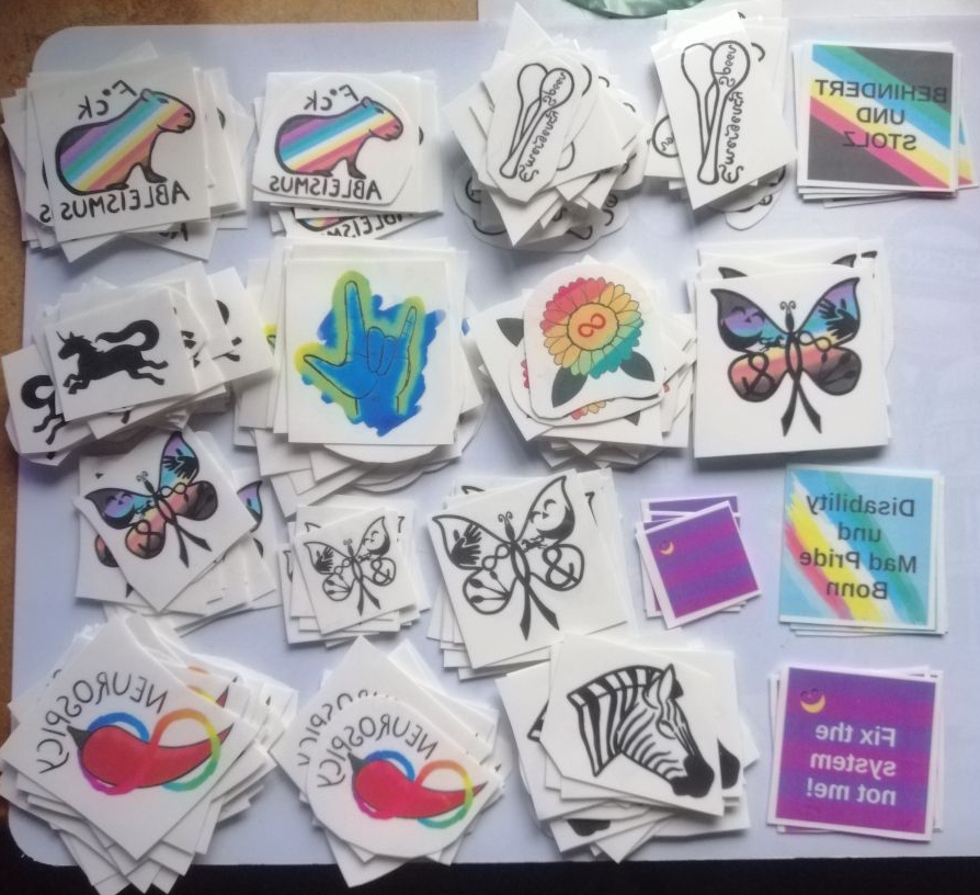Einmal-Tattoo Motive: F*ck Ableismus Capybara, Autistic Pride Sonnenblume, I love you Gebärde in D*deaf Pride Farben, Emergency Spoons, CIMND* Schmetterling, Neurospicy Chilli, Zebra, Einhorn. Und von 2024: Behindert und Stolz mit einer Pride Fahne, Disability und Mad Pride Bonn Logo, Mad Pride Fahne, Fix the system, not me! mit Fahne.