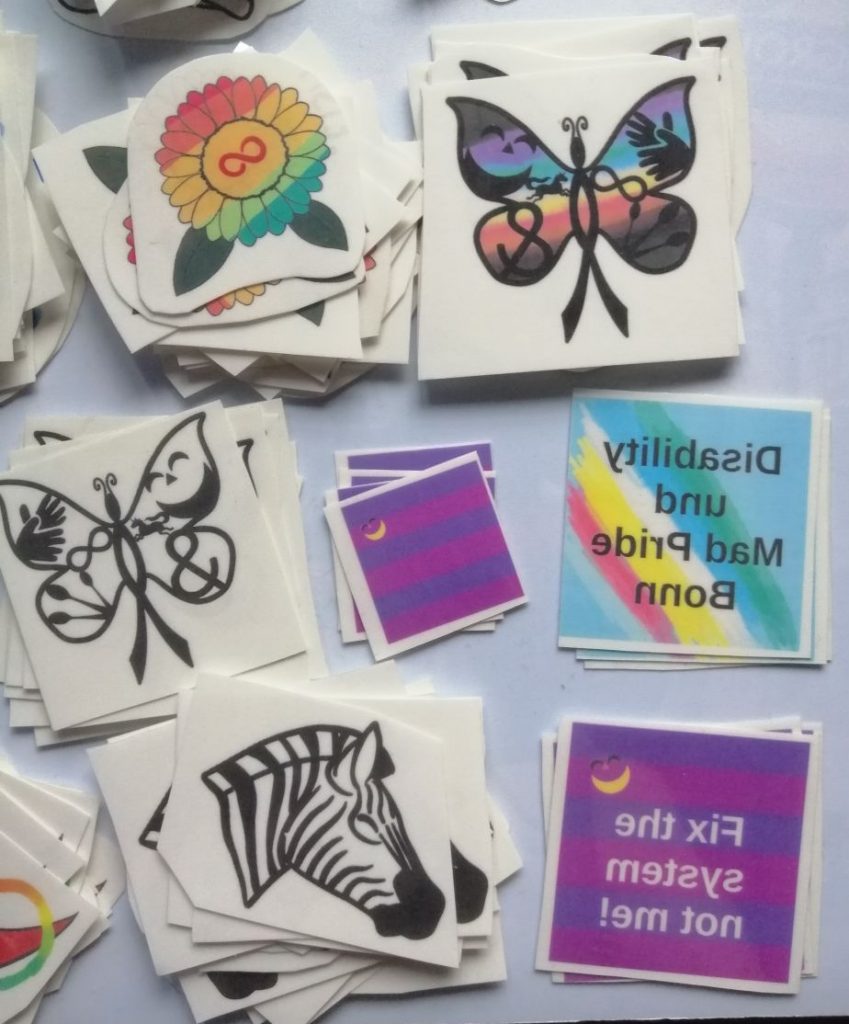 Einmal-Tattoo Motive: Autistic Pride Sonnenblume, CIMND* Schmetterling, Zebra. Und von 2024: Disability und Mad Pride Bonn Logo, Mad Pride Fahne, Fix the system, not me! mit Fahne.