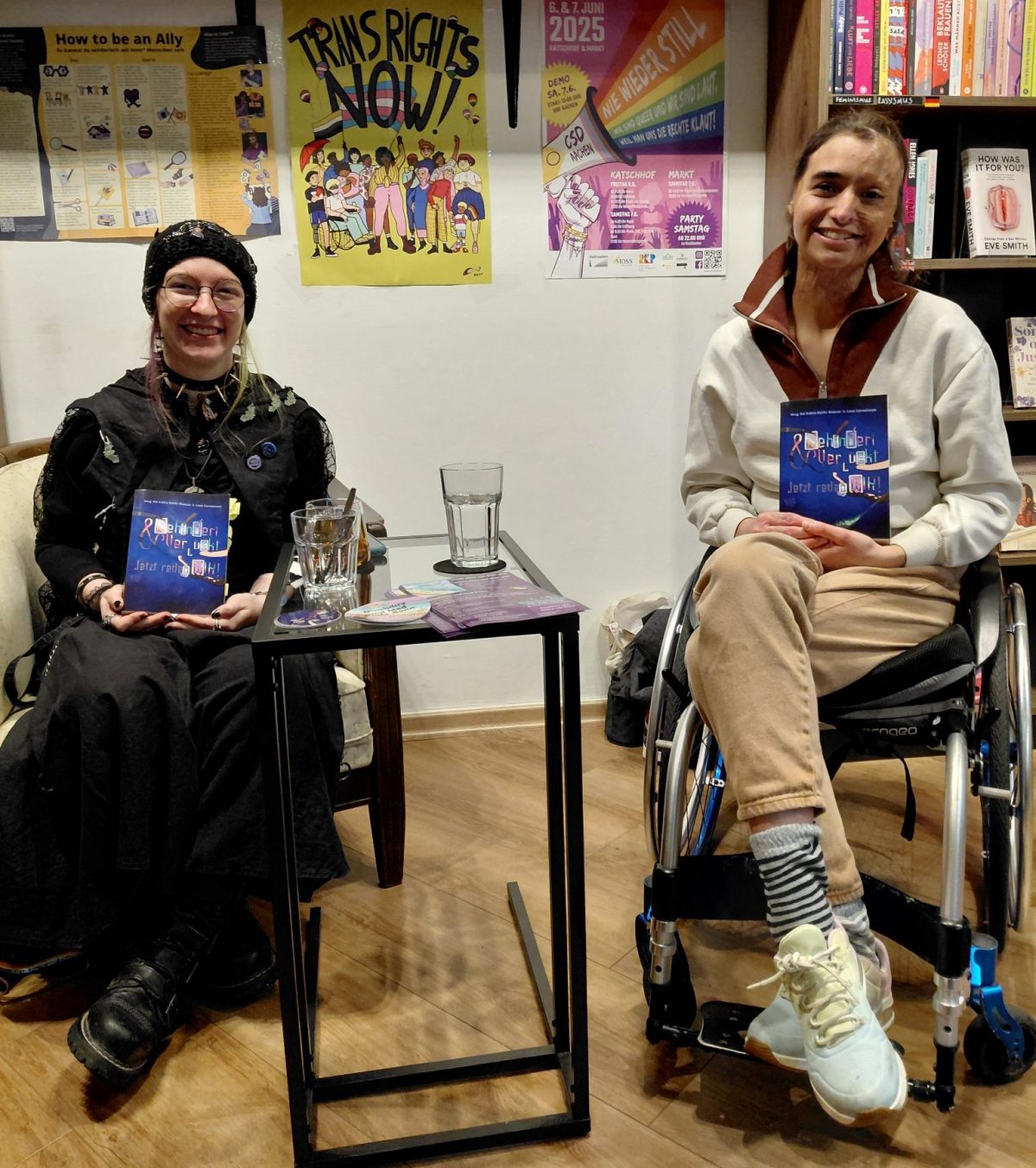 Auf dem Foto sind Koi und Lee. Beide haben das Buch: "Behindert & Verrückt - Jetzt reden Wir!" in der Hand. Koi sitzt auf einem Sessel. Lee sitzt in einem Rollstuhl. Koi hat schwarze Klamotten an und eine schwarze Mütze. Lee hat weiße und braune Klamotten an. Beide lächeln.