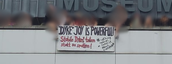 Es sind Personen und ein Schild zu sehen. Die Personen sind alle verschwommen. Auf dem Schild steht: Dyke* joy is powerful! Stabile Dykes* teilen statt zu spalten! #nothing radical or feminist about transphobia. Das ist teils Englisch und heißt etwa: Dyke* Freude ist stark! Stabile Dykes* teilen statt zu spalten! # Nichts ist radikal oder feministisch an Trans*feindlichkeit.